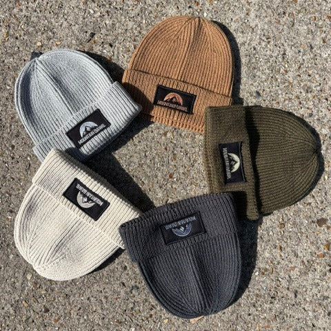 Five ribbed knit beanie hats arranged in a circle on a textured concrete surface. The hats are light gray, tan, off-white, and dark charcoal gray, each featuring a black rectangular patch on the cuff with a white stylized Mountain Dawg logo