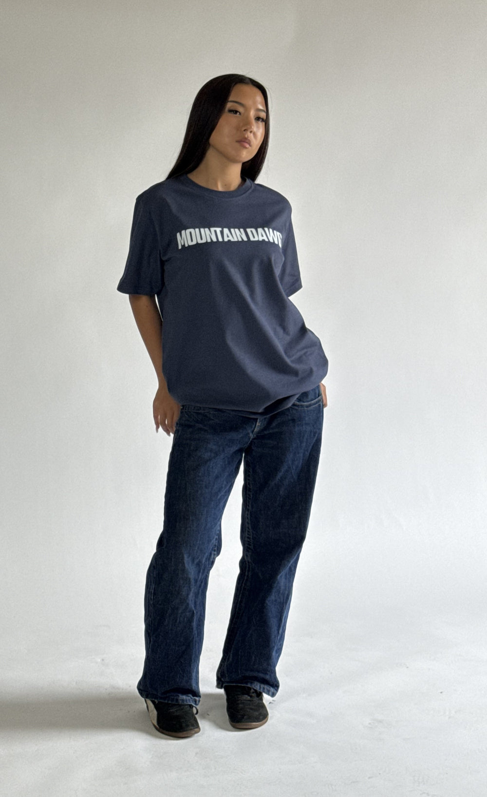 A model wearing a dark navy blue organci cotton t-shirt with 'MOUNTAIN DAWG' printed in white across the chest, paired with dark blue jeans and black shoes, standing against a plain light gray studio background.