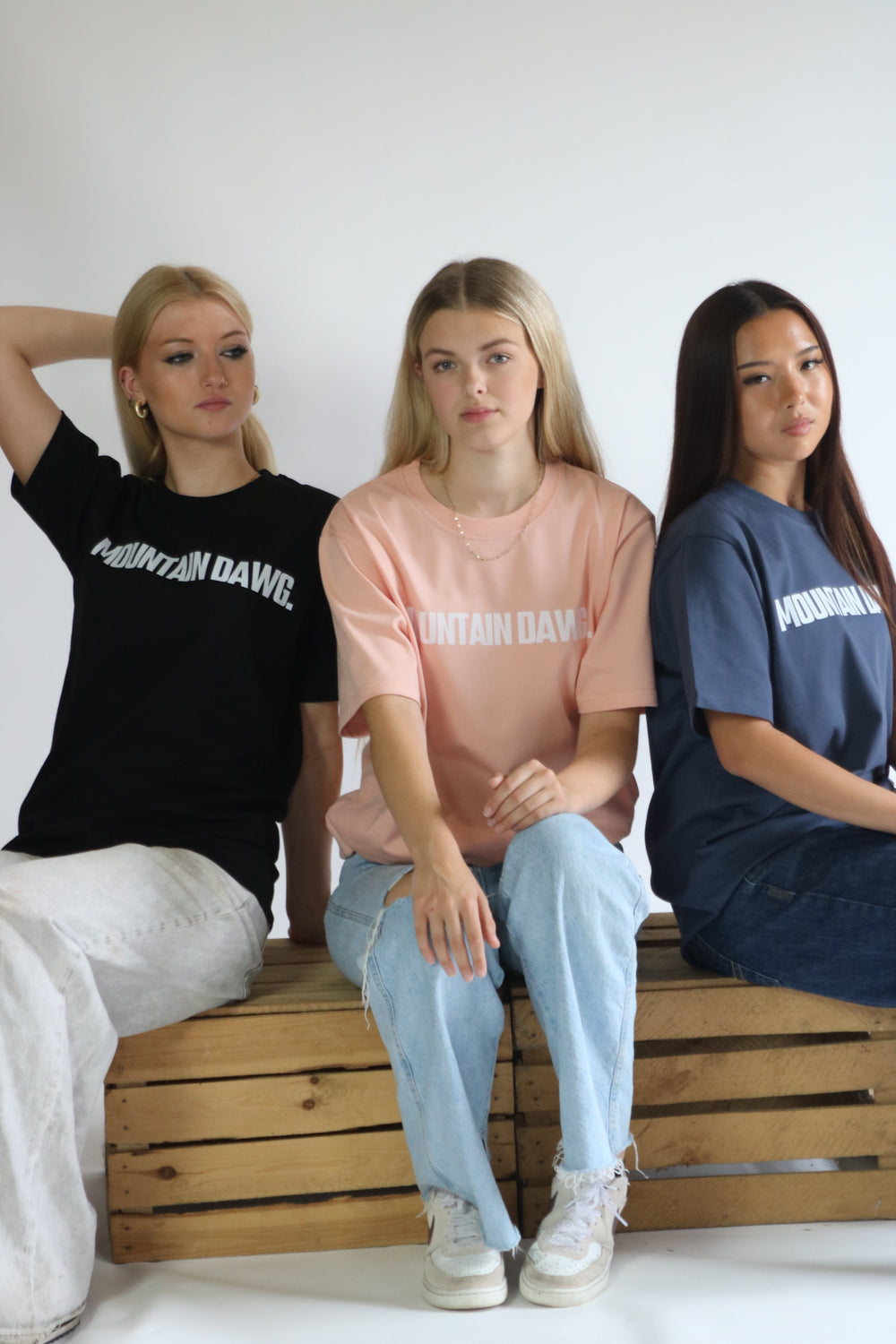 Three young women sitting on a wooden crate bench, wearing organic cotton mountain dawg t-shirts with 'MOUNTAIN DAWG' printed in white across the chest.