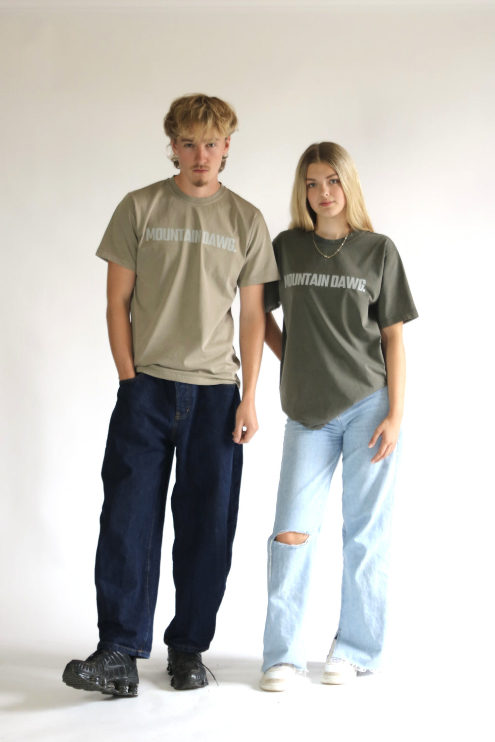 Two models standing against a white studio background wearing organic cotton mountain dawg t-shirts with 'MOUNTAIN DAWG' printed across the chest. The model on the left wears a light beige t-shirt and dark blue jeans, while the model on the right wears an olive green t-shirt and light blue ripped jeans.