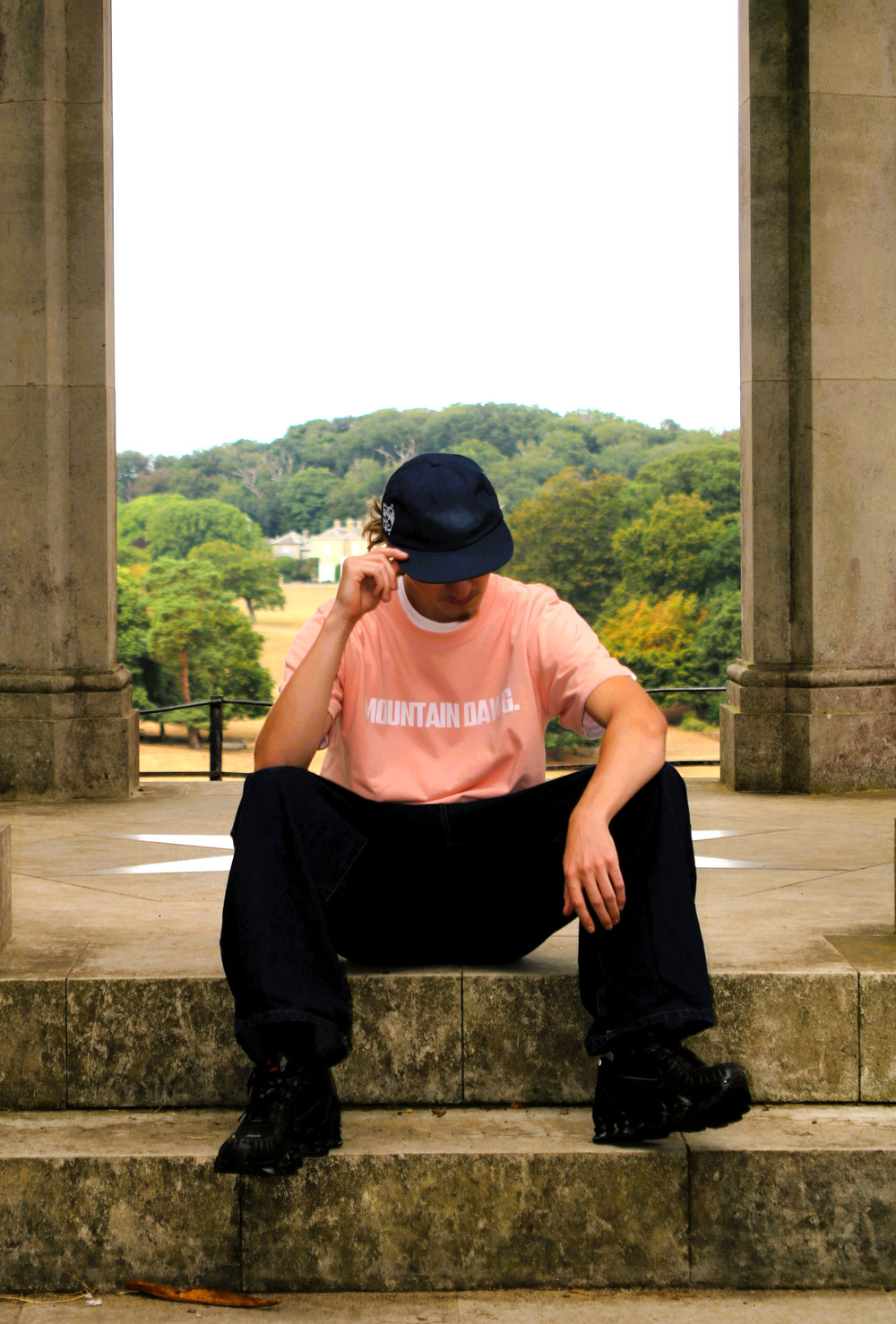 A person sitting on stone steps between columns, wearing a dusty pink organic cotton t-shirt with 'MOUNTAIN DAWG' printed on the front, black pants, black boots, and a dark navy cap, with a park in the background.