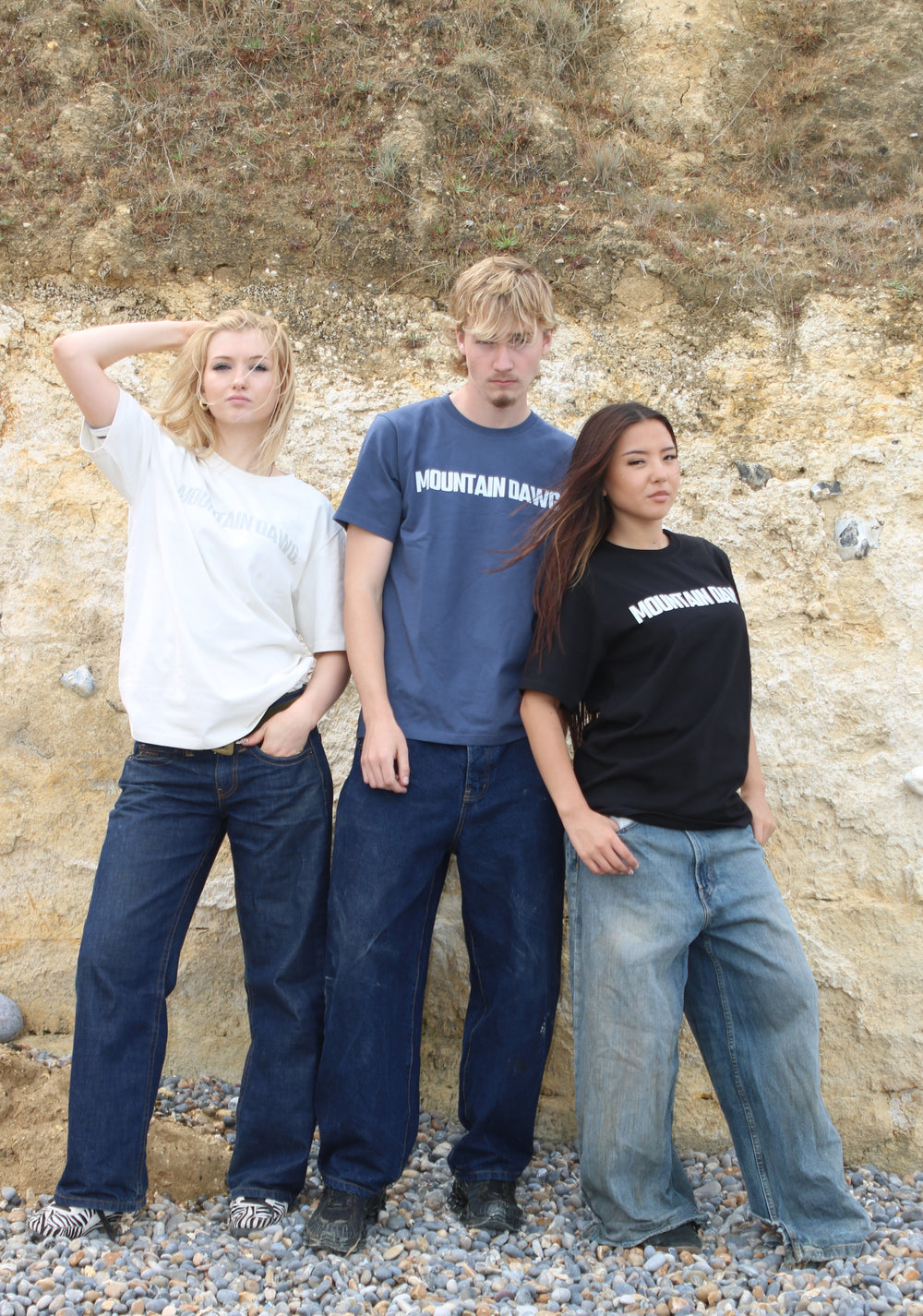 Three young adults standing together outdoors against a rocky, earthy cliff face. The person on the left wears a white mountain dawg organic cotton T-shirt and dark blue jeans.