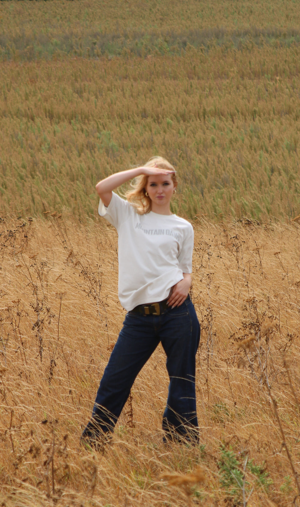 A woman stands in a dry grass field wearing a vintage white organic cotton mountain dawg t-shirt with faint text on the chest and dark blue jeans, looking towards the camera while shielding her eyes with her hand.