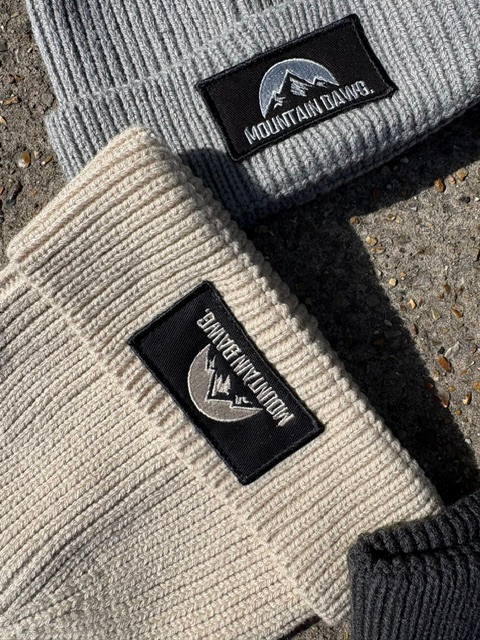 Close-up of three ribbed knit beanie hats laid on a rough outdoor surface. The beanies are light gray, oatmeal, and graphite gray, each featuring a black rectangular patch with a mountain logo and the text 'MOUNTAIN DAWG'.