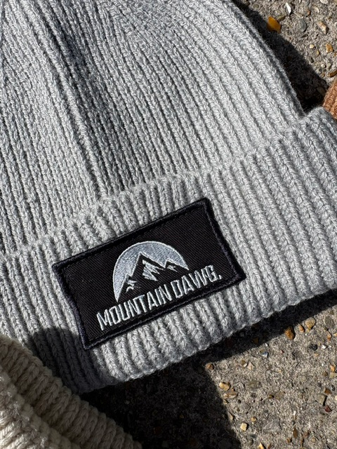 A close-up of a light gray ribbed knit beanie with a black rectangular embroidered patch on the cuff. The patch features a mountain graphic and the text 'MOUNTAIN DAWG.