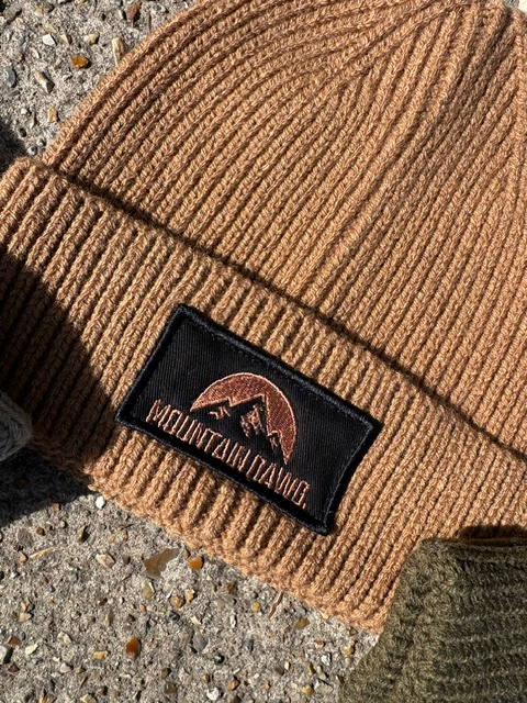 A close-up of a ribbed biscuit colour beanie with a black embroidered patch reading 'MOUNTAIN DAWG' and a mountain graphic, laid on a rough concrete surface.
