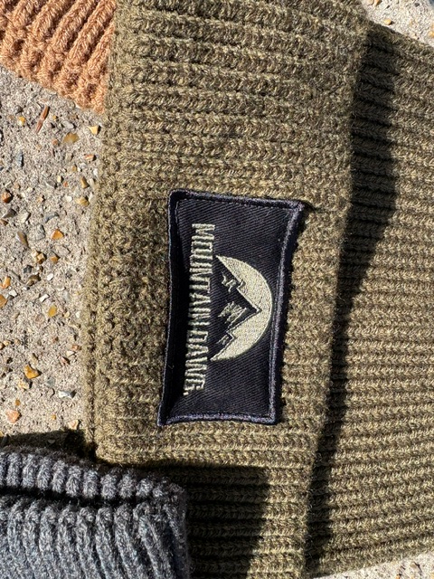 Close-up of a ribbed knit beanie in military green with a black rectangular patch that reads Mountain Dawg with a mountain graphic.