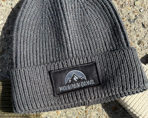 Close-up of a ribbed knit beanie cuff with a black rectangular patch reading MOUNTAIN DAWG and a mountain logo, shown alongside other color variants of the same hat on a concrete surface.